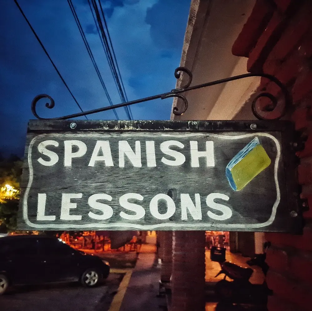 A wooden sign asvertsigin 'Spanish lessons' hanging on a metal awning outside a building.