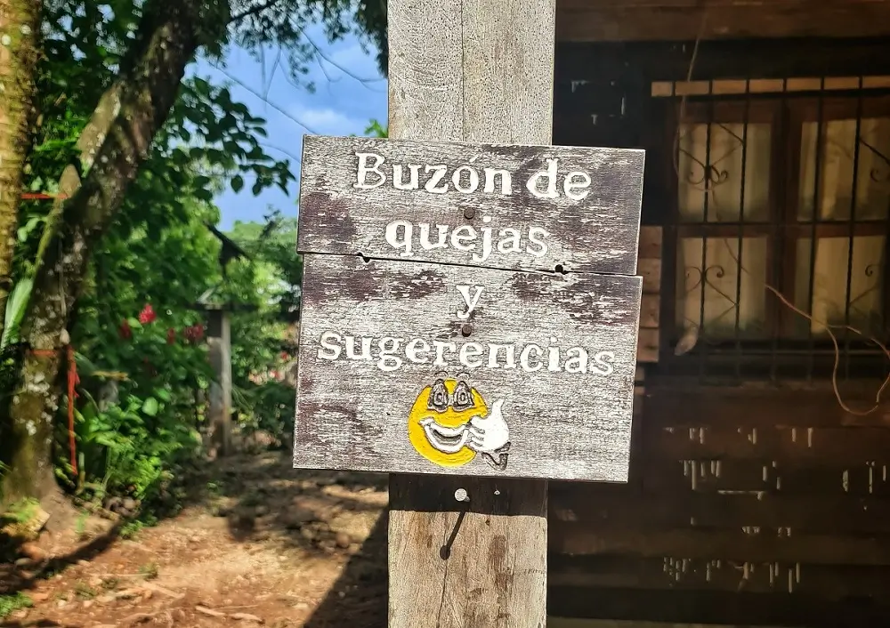 A wooden sign outside a building in Mexico with writting in Spanish which translates to 'Complaint mailbox and suggestions" with an emoji of a character with a smiling face and giving a thumbs up.