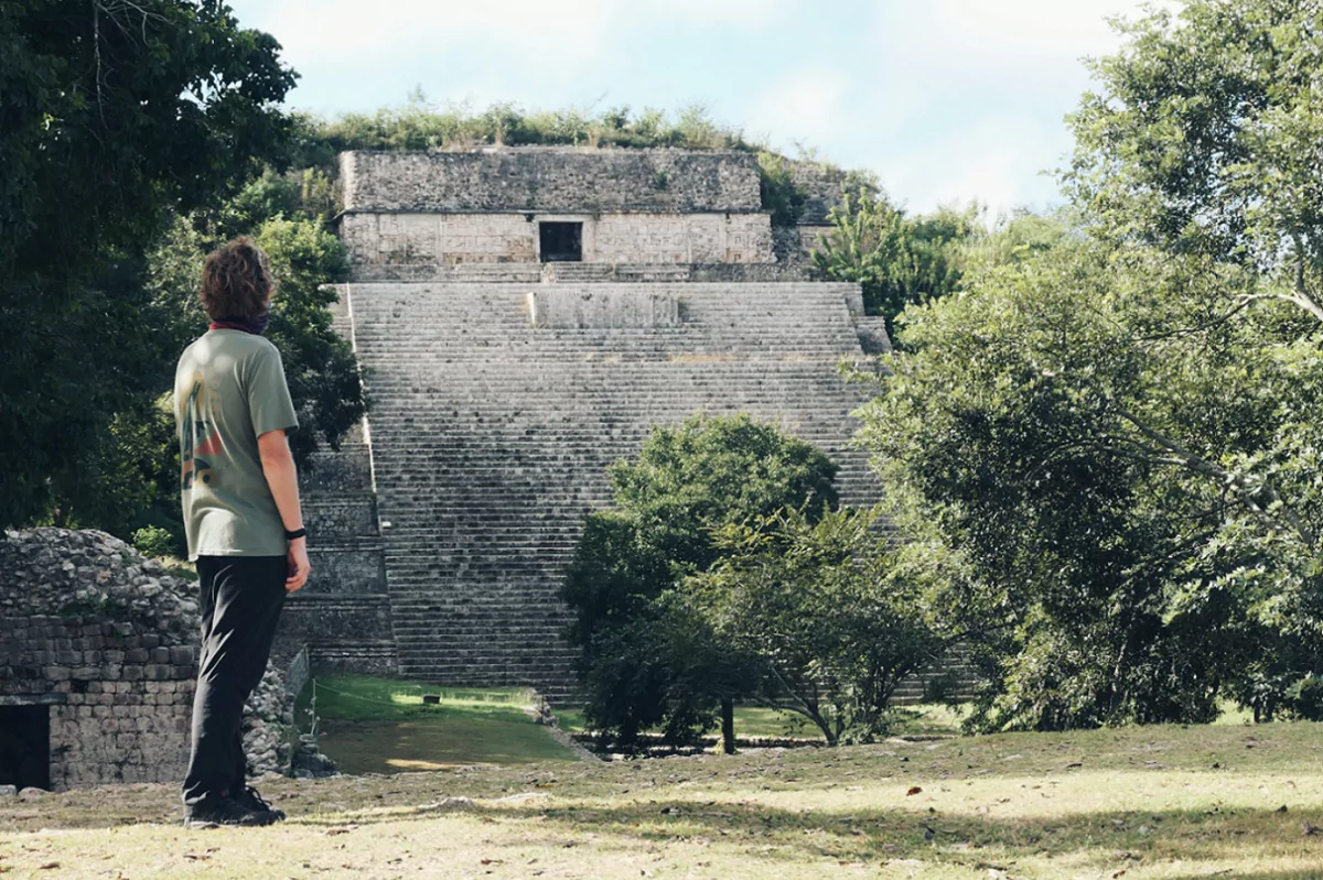 A person looking at a Mayan temple ruin within Uxmal, surrounded by jungle trees.