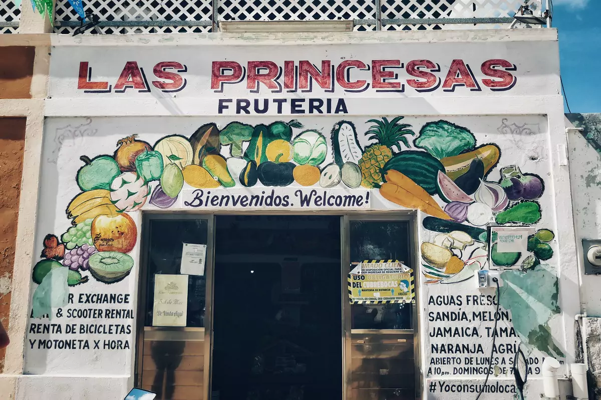 The shop front of a fruit and vegetable store in Progresso, near Merida, with a mural on the front of a multitude of fruits and vegetables with writing on the wall by the entrance of fruit juices for sale.