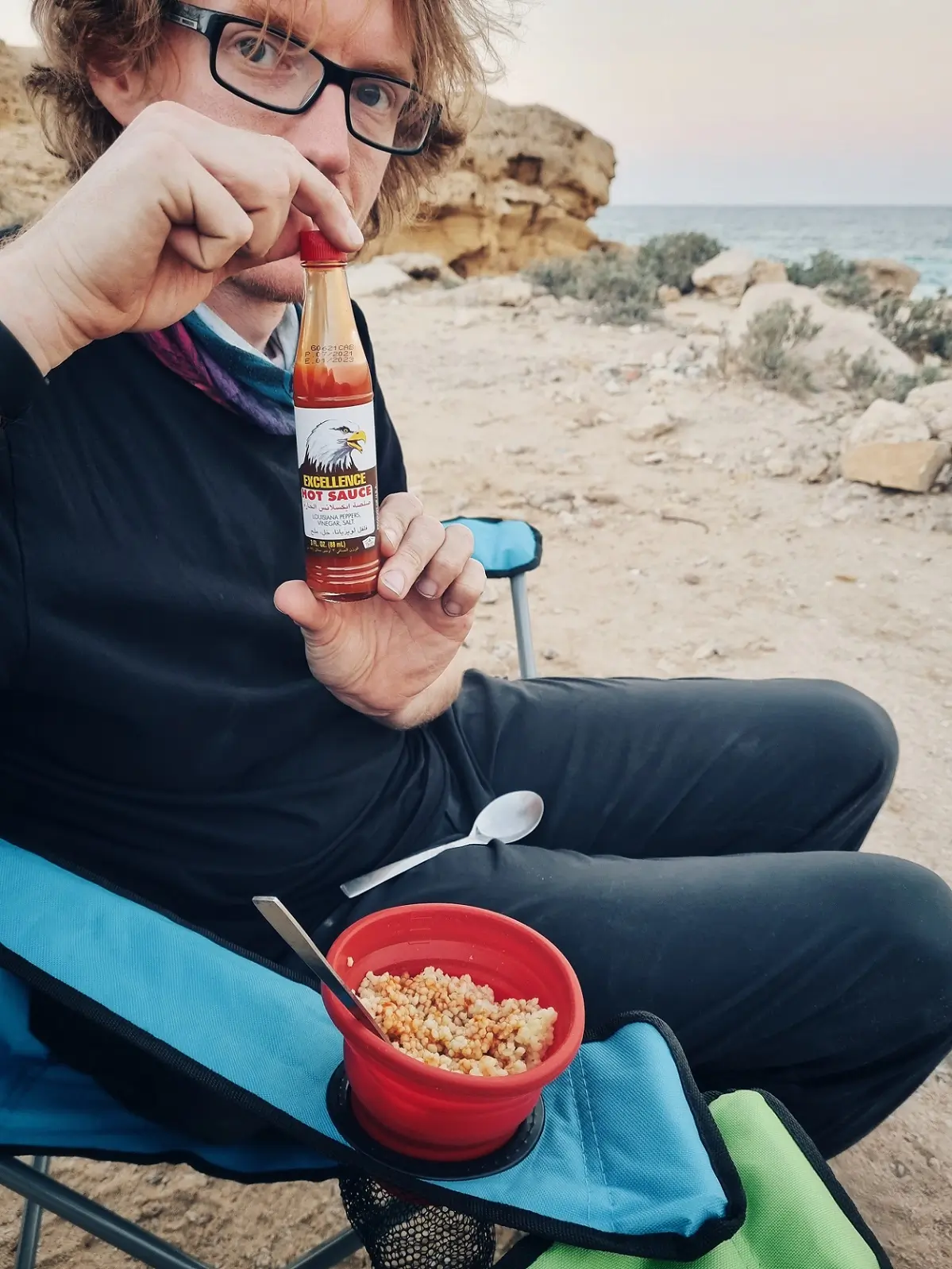 A person sat on a chair at a campsite with a bottle of hot sauce for their cous cous dinner.