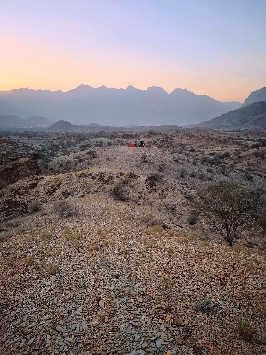 View of the expansive, sparse West Hajar mountains in Oman with a white 4x4 and campsite in the distance.