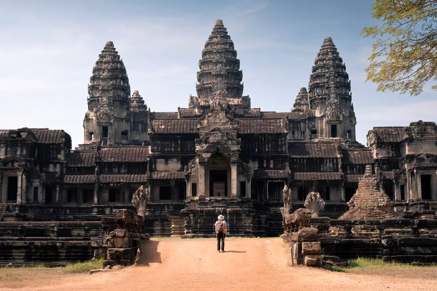 A person wearing a yellow and grey backpack, standing on a dusty path before the truly impressive three-towered stone temple at Angkor Wat.