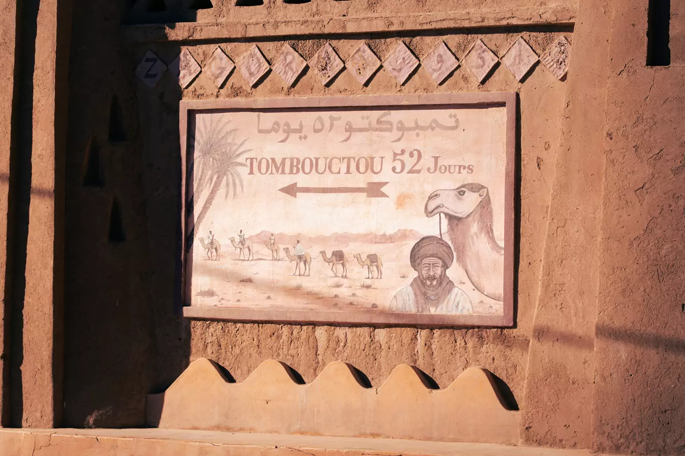 A sign featuring camels and Arabic riders, indicating it is '52 days journey to Tombouctou'.