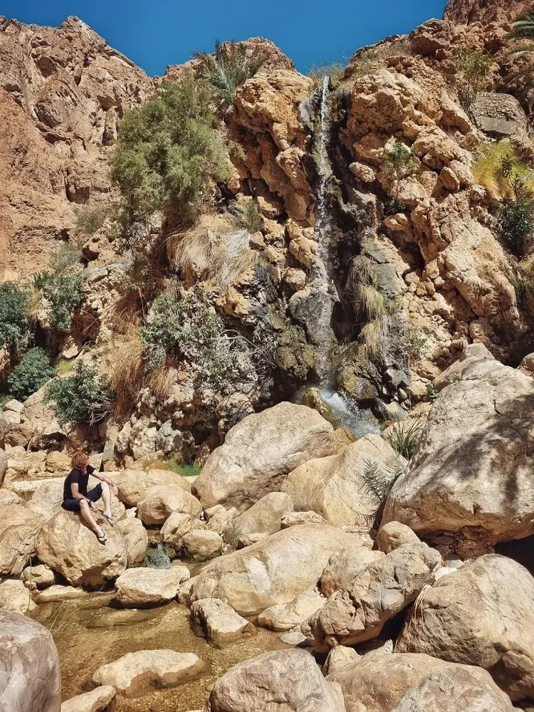 A waterfall in the canyon of Wadi Shab with a person sat on a boulder below.