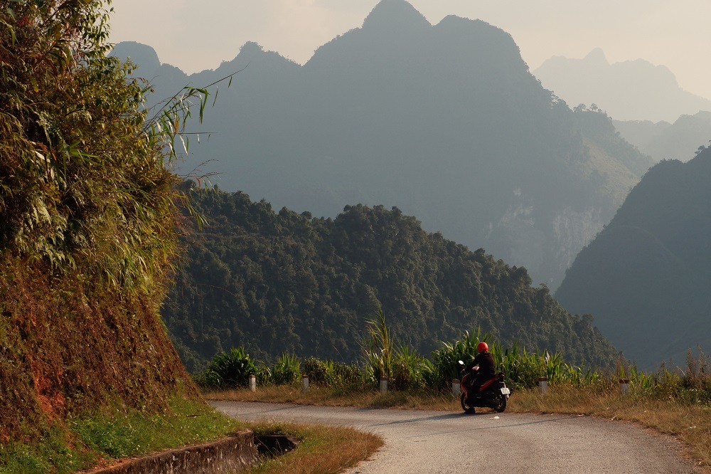 A motorcyclist with a red helmet by the side of a winding road leading through the mountains of northern Vietnam.