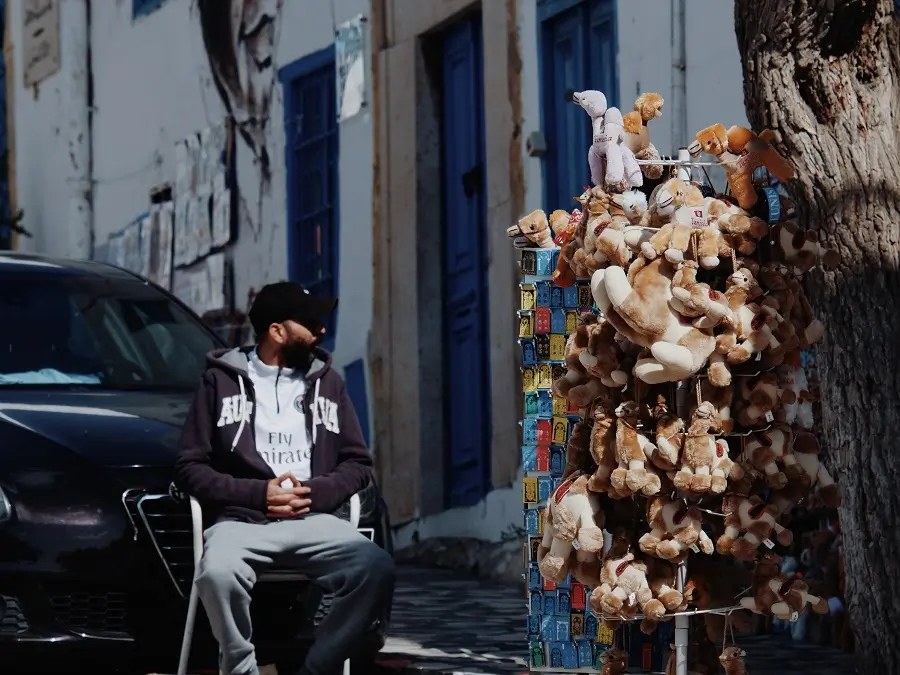 A merchant sat on a chair beside souvenirs of stuffed animal toys and magnets.