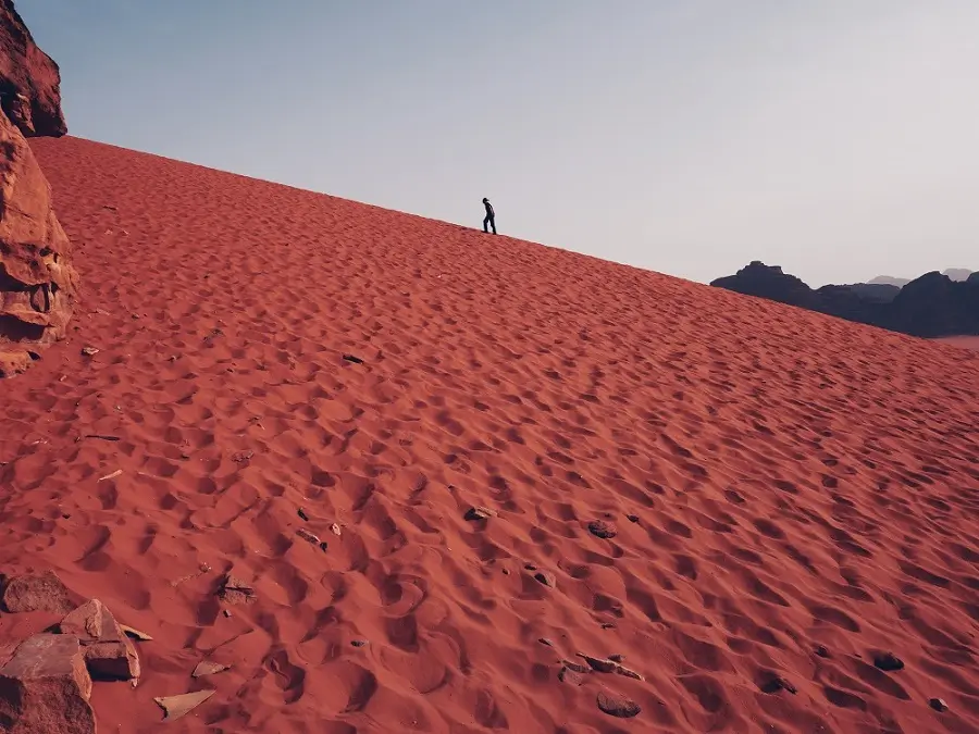 A person walking along a steep, deep red sand dune in Wadi Rum, Jordan.