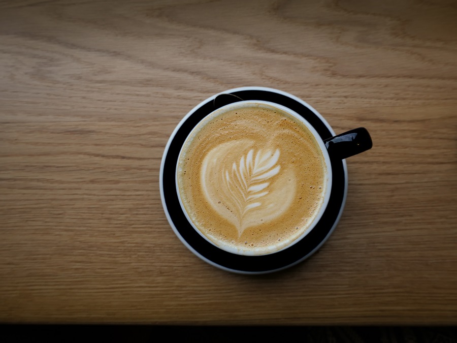A cup of coffee, sat on a black saucer, with a floral decoration in the foam.