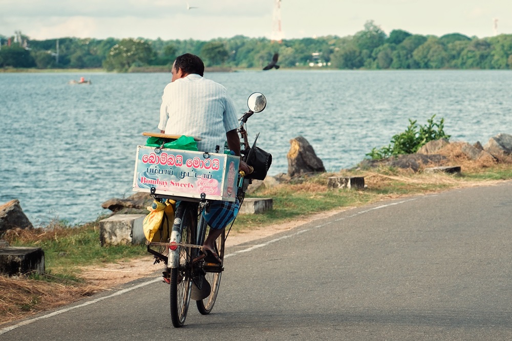 A cyclist-vendor on a quiet road by a lake in Polonnaruwa, selling sweets in Sri Lanka.