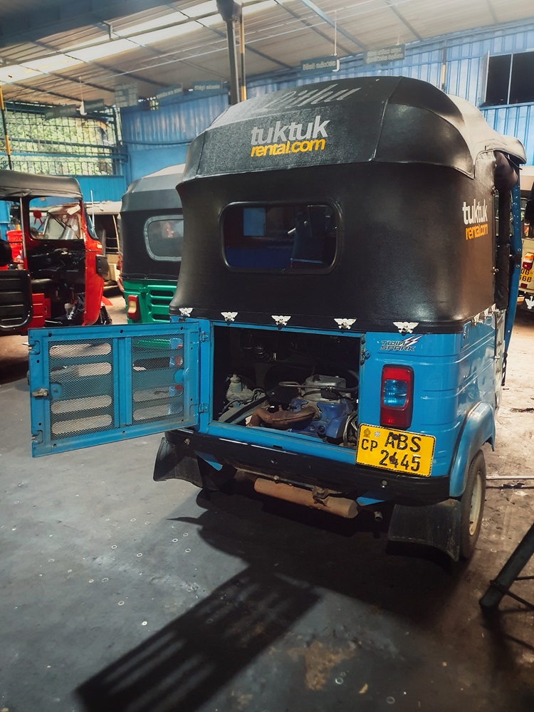The rear of a blue rental tuktuk in a Sri Lankan mechanic shop with the engine compartment open.