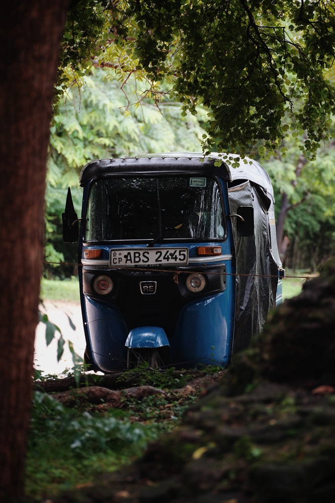 A blue Sri Lanka rental tuktuk with the rain flaps down, parked under a tree.