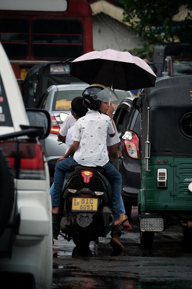 A motorcycle driving in the rain in Sri Lanka with the passenger holding up an umbrella to keep them dry.