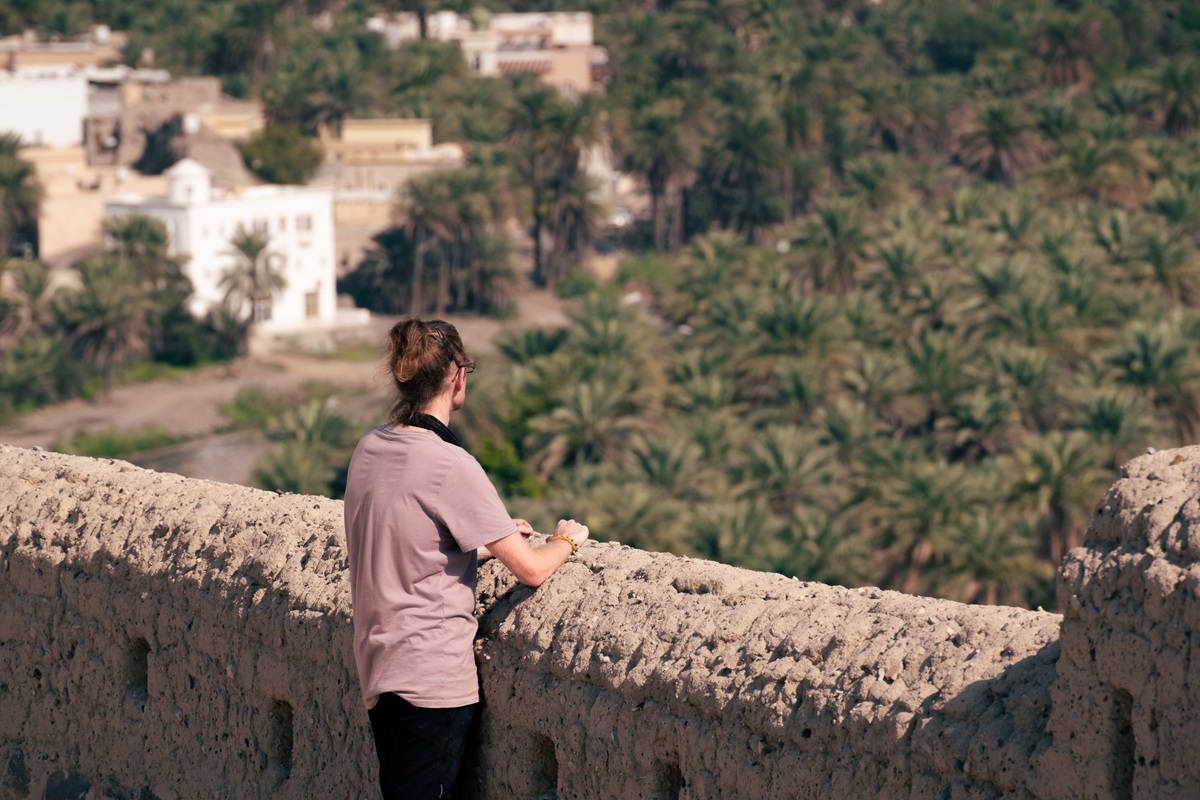 A person looking over mudbrick walls at the palm trees and houses of Samail village below.