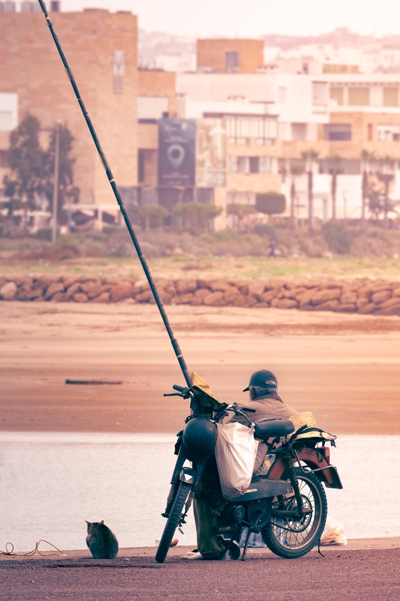 A man fishing in the river, by their motorbike, at Rabat with a cat sat nearby.