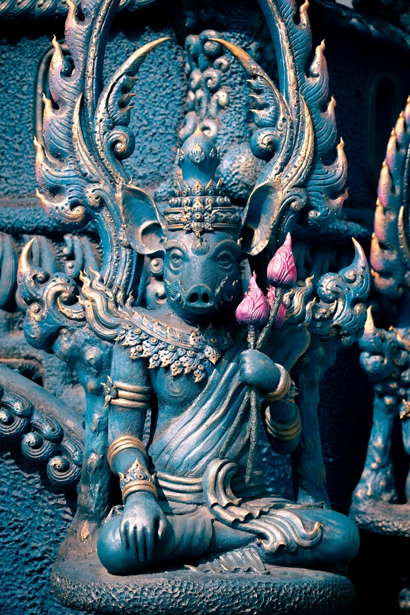 A part pig, part human statue sitting down and holding a lotus flower at the Blue Temple in Chiang Rai.