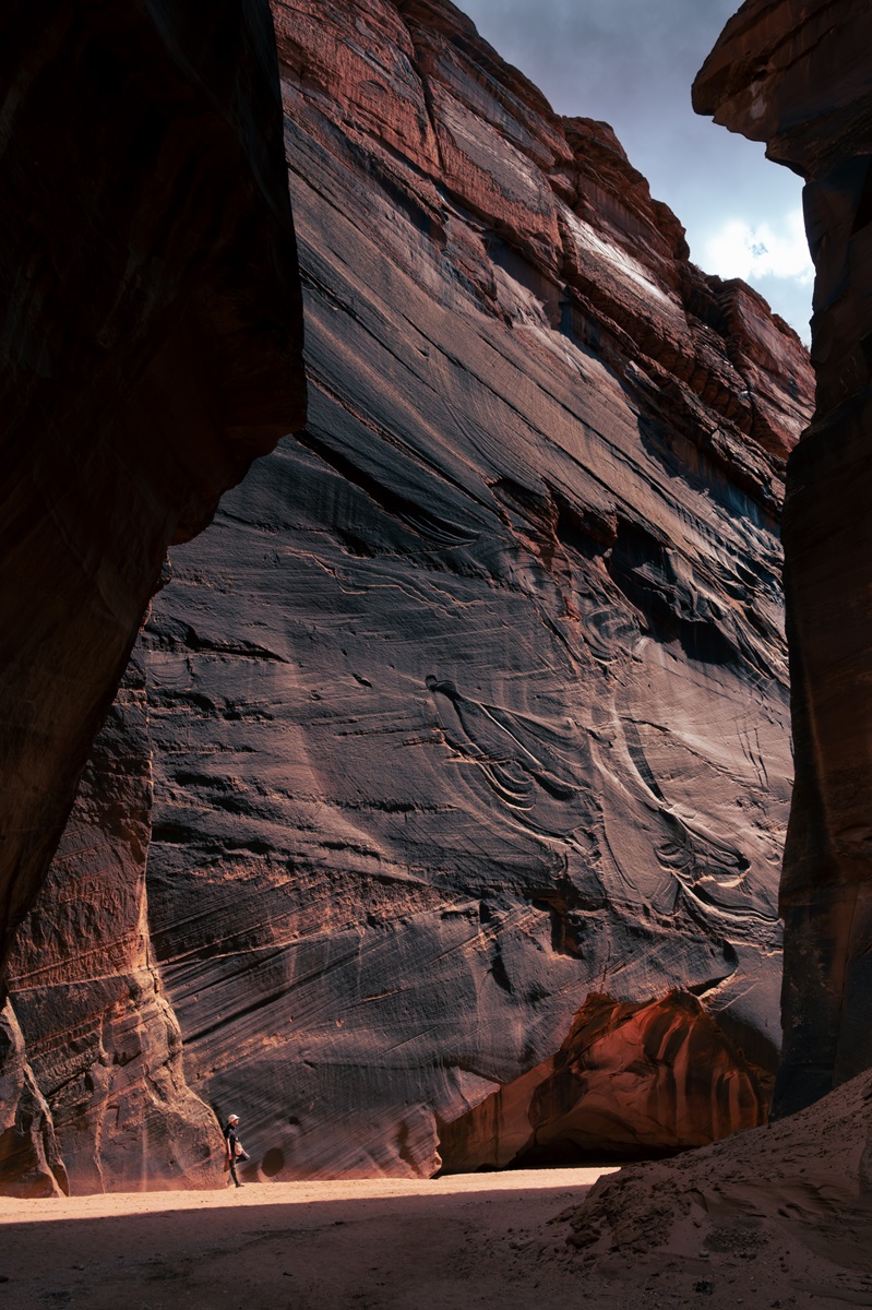 A hiker leaning against the base of a towering cliff wall of a slot canyon in the sunlight.
