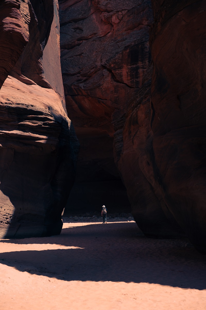 A hiker wearing a white hat walking along the sandy bottom of a slot canyon with towering sandstone walls curving around them.