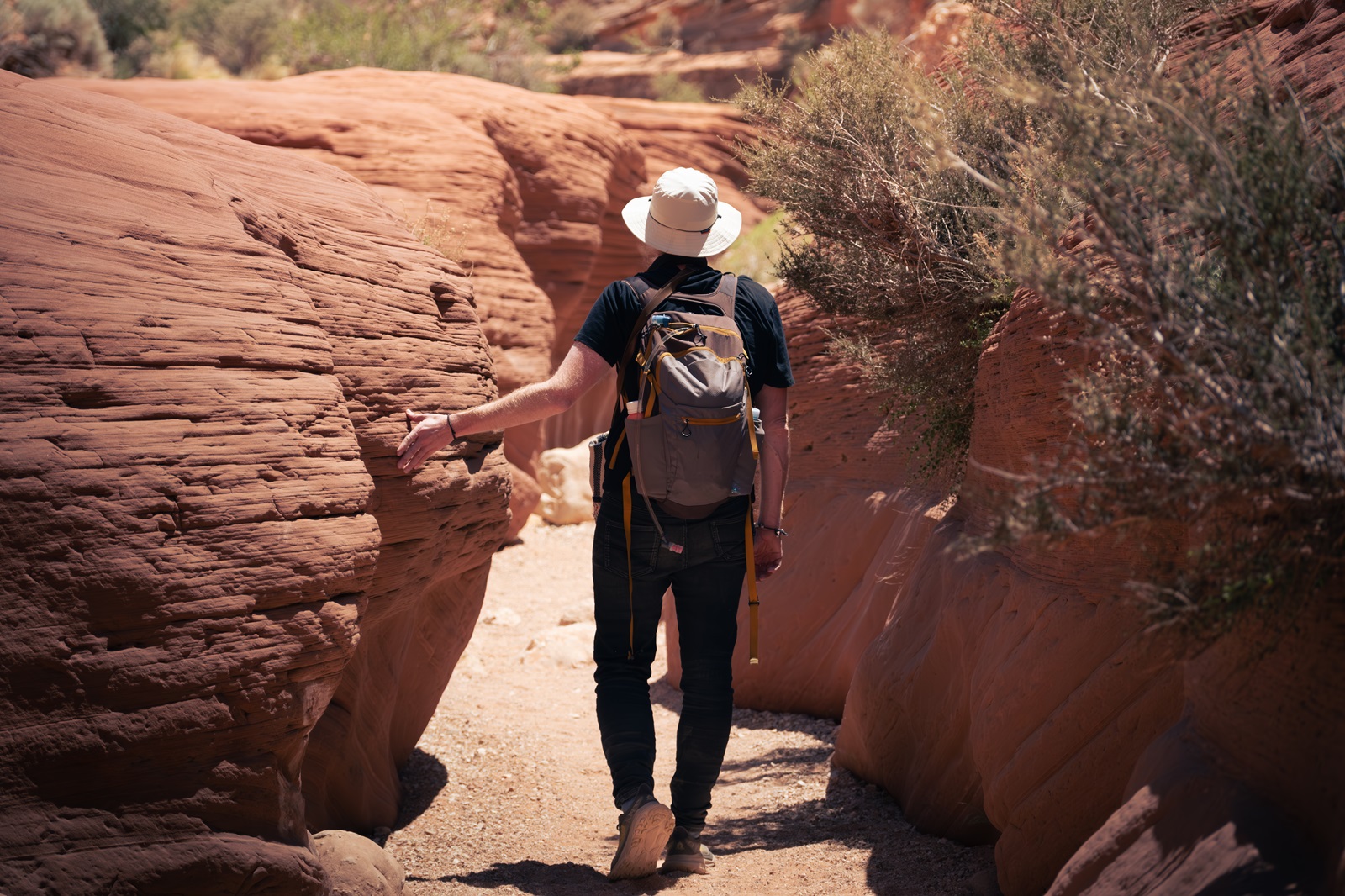 A hiker with their hand on a sandstone rock formation as they walk along a narrow path.