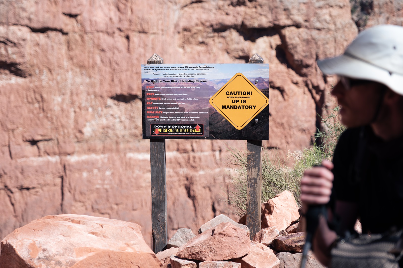 A hiker beside a caution sign indicating the dangers of hiking the South Rim trails of the Grand Canyon.
