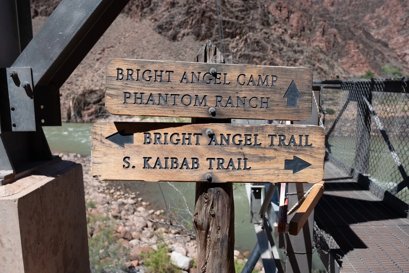 Wooden trail signs by the Colorado River in the Grand Canyon pointing to "Bright Angel Camp, Phantom Ranch" and "Bright Angel Trail, S. Kaibab Trail."