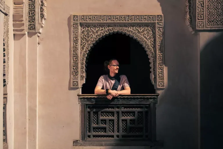 A person standing at a window with arms on a wooden ledge, looking to their left across an atrium in a madrasa.