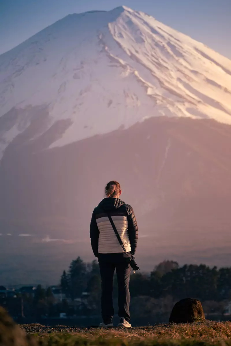 A photographer standing in a grassy field with the snow peaked Mount Fuji in the distance.