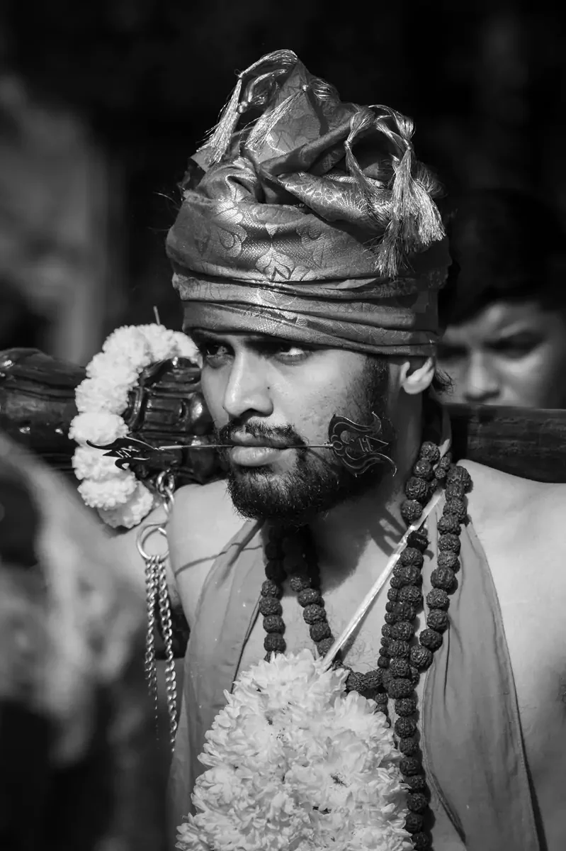 A person with beads and a flower garland around their neck with a spear through their tongue, at Thaipusam.