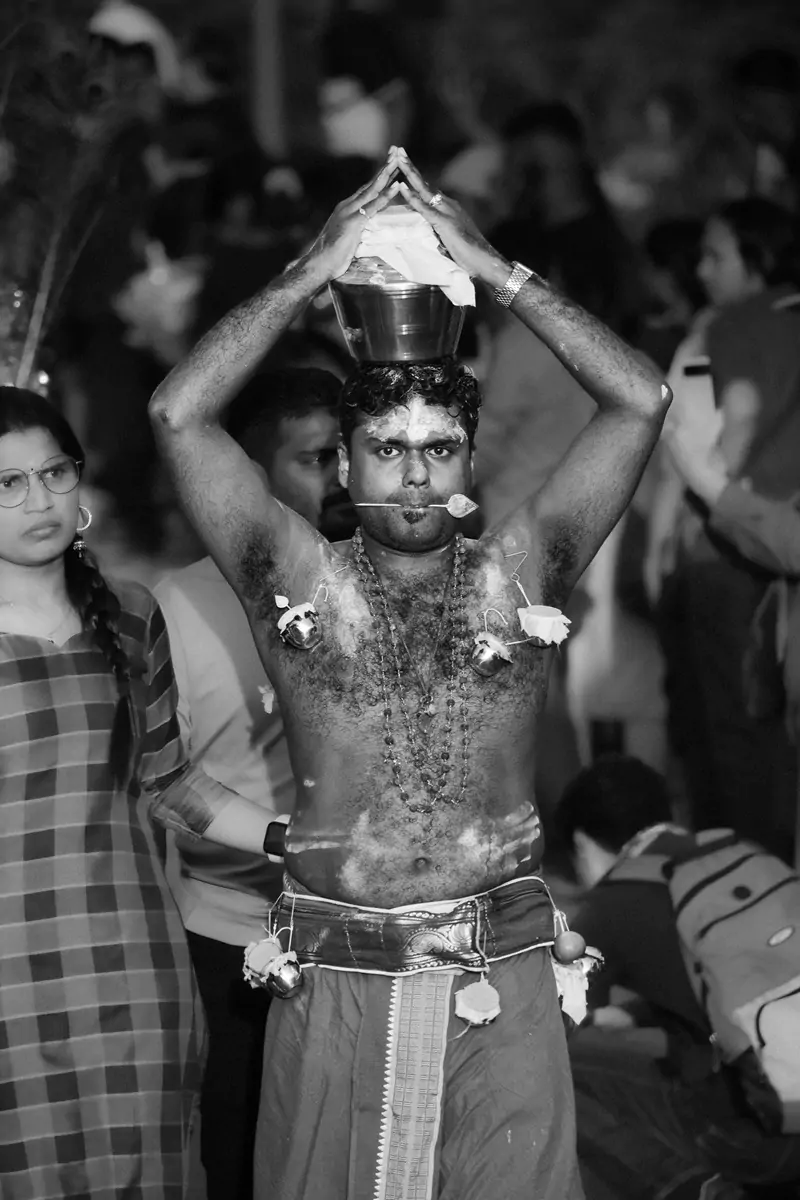 A person with a spear through their cheek holding a silver milk pot atop their head at Thaipusam.