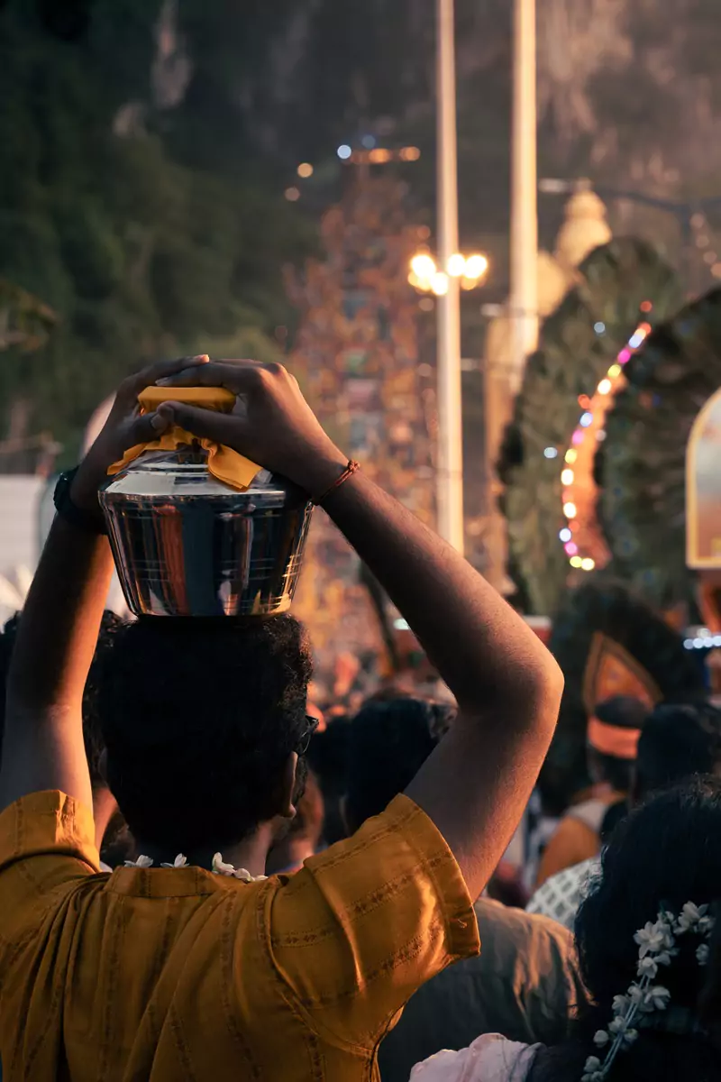 A person wearing yellow, the favoured colour of Lord Murugan, and holding a silver milk pot on their head using two hands.