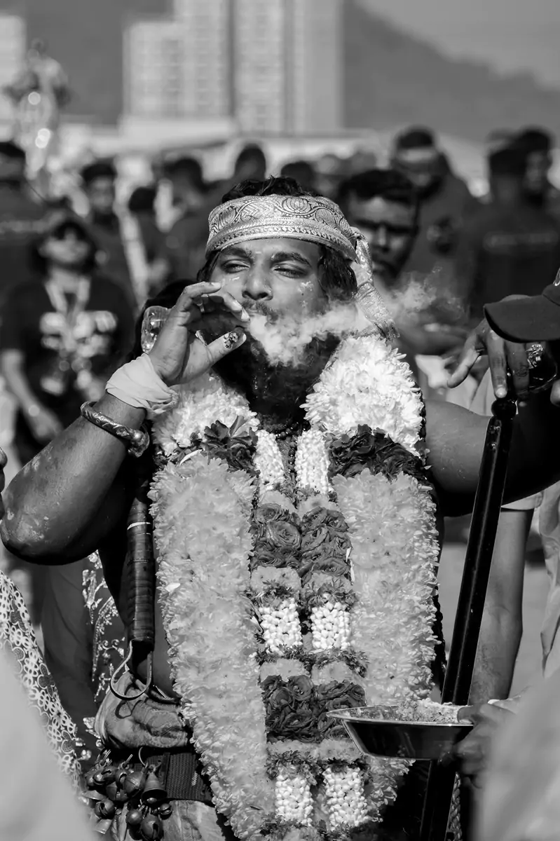 A person smoking a cigar and puffing out smoke, wearing a colourful garland of flowers.