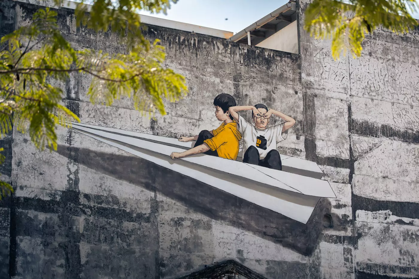 A fresco of two boys sat inside a large paper plane, painting on a building in Ipoh, Malaysia, by Ernest Zacharevic.