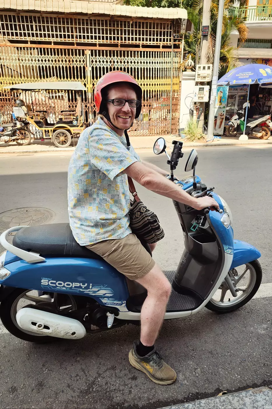 A smiling motorcyclist parked on the side of a road in Siem Reap wearing a red motorcycle helmet.