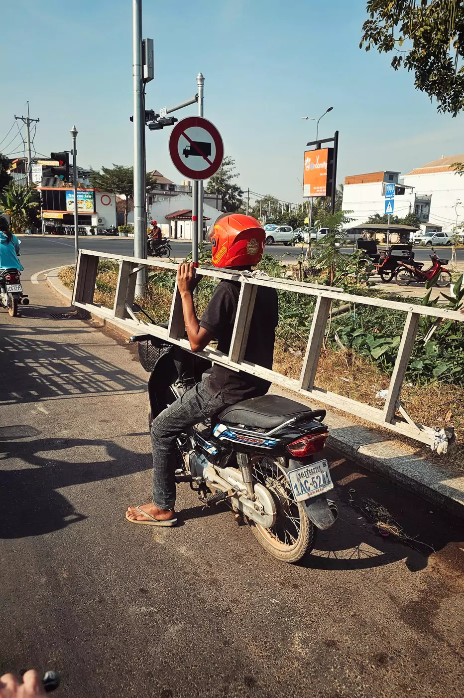 A motorcyclist waiting at a junction in Siem Reap, carrying a metal ladder over one shoulder.