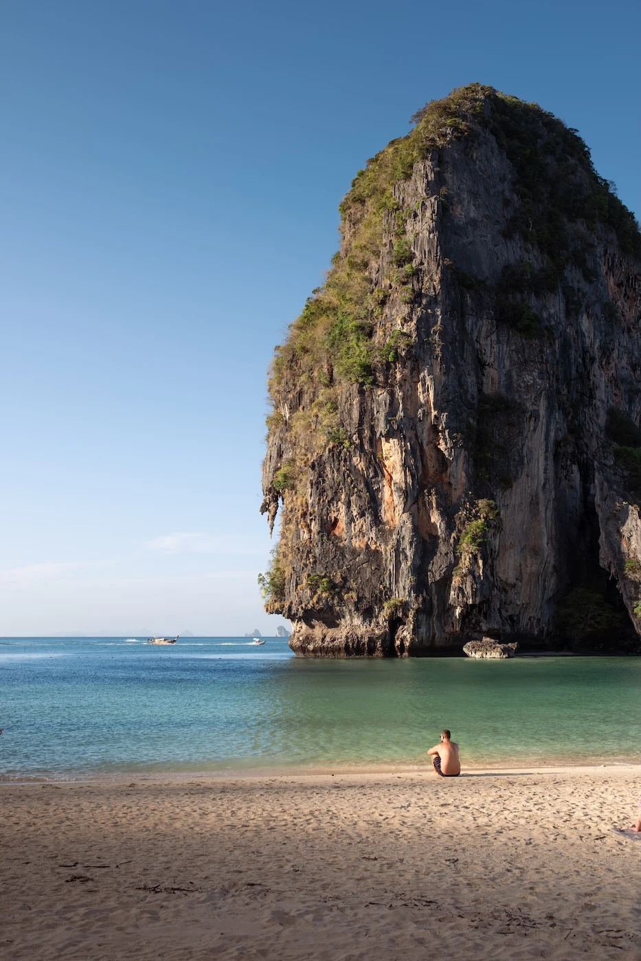 A tourist sat alone a stretch of sand at Phra Nang Beach with a large karst rock structure in the sea, on Railay, Thailand
