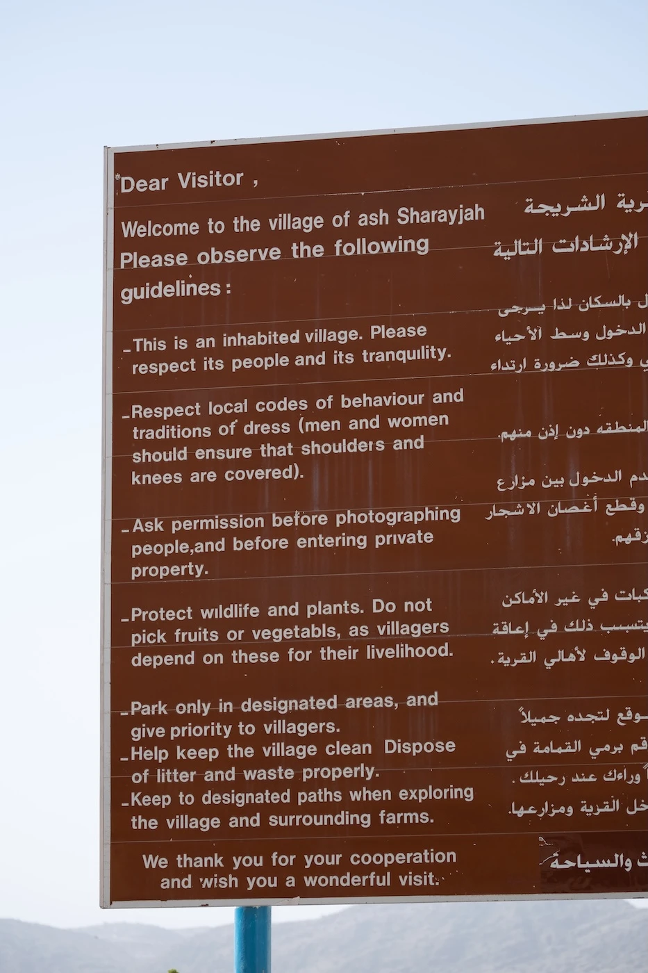 A large brown metal sign at the village of Ash Sharayjah, written in English and Arabic, with rules for visitors to the village.