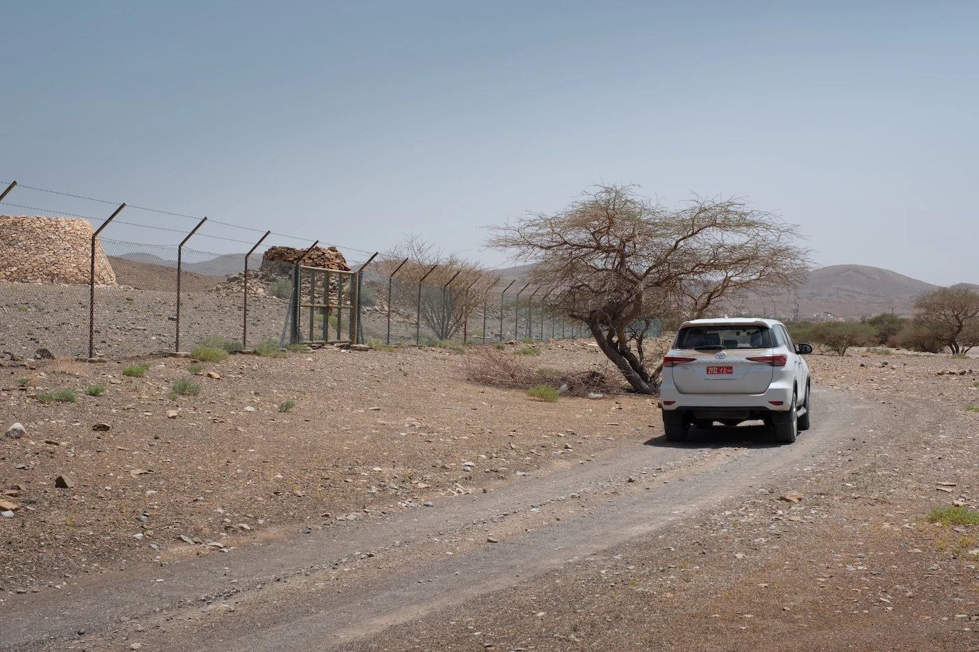 A parked off-road vehicle next to a locked metal gate at the arid Bat Archaeological Site in Oman.