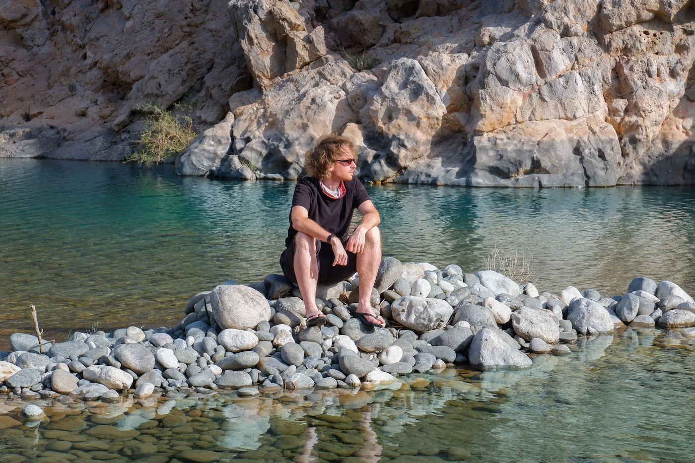 A traveller sitting on a pile of rocks within a blue pool of water at Wadi Dayqah.