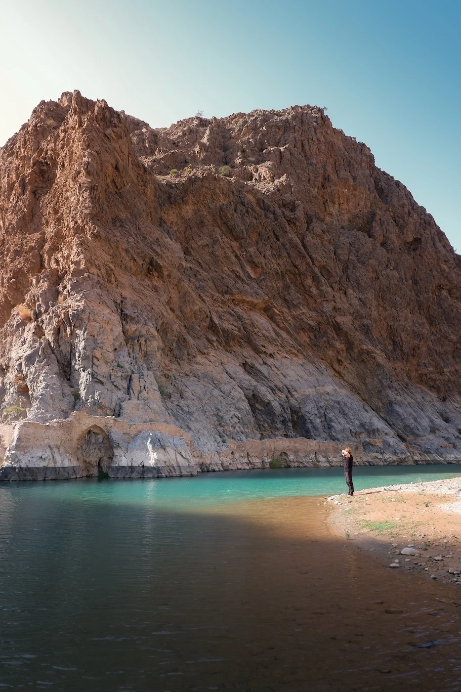 A person standing by the shoreline of a pool of water, edged by a rocky outcrop on the opposite side within Wadi Dayqah.