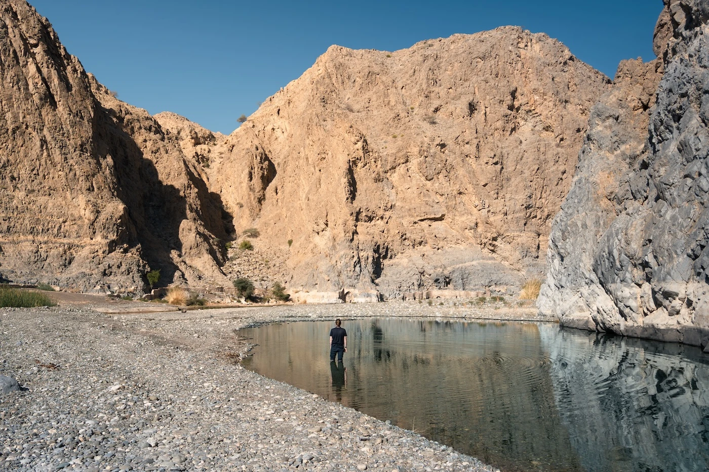 A person standing in clear waters up to their knees, with reflections of the surrounding valley on the surface of the water at Wadi Dayqah near Muscat, Oman.