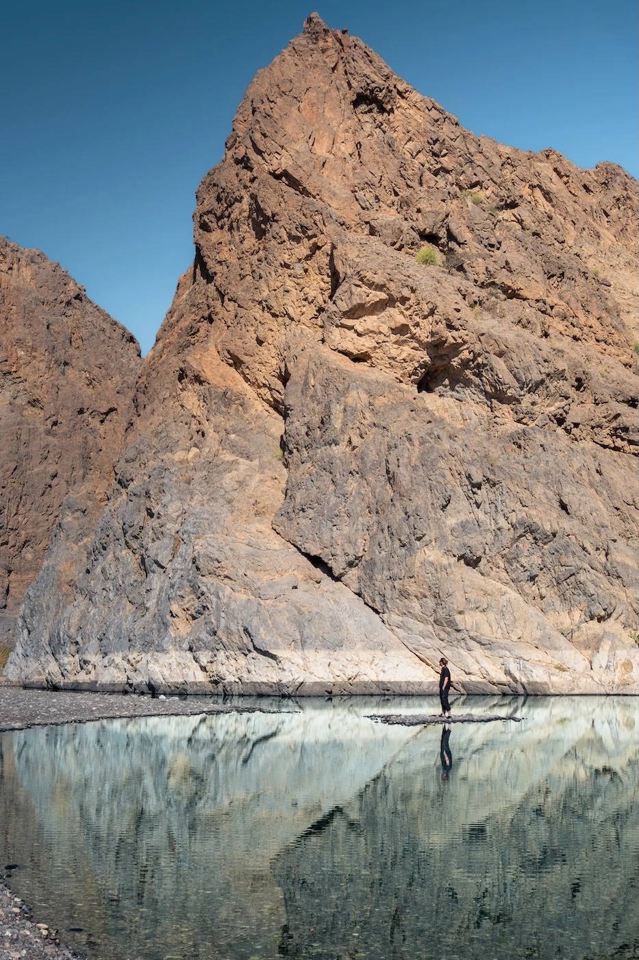 A person standing in a pool of calm, clear water with the rocky backdrop of the surrounding Wadi Dayqah reflecting in the waters.