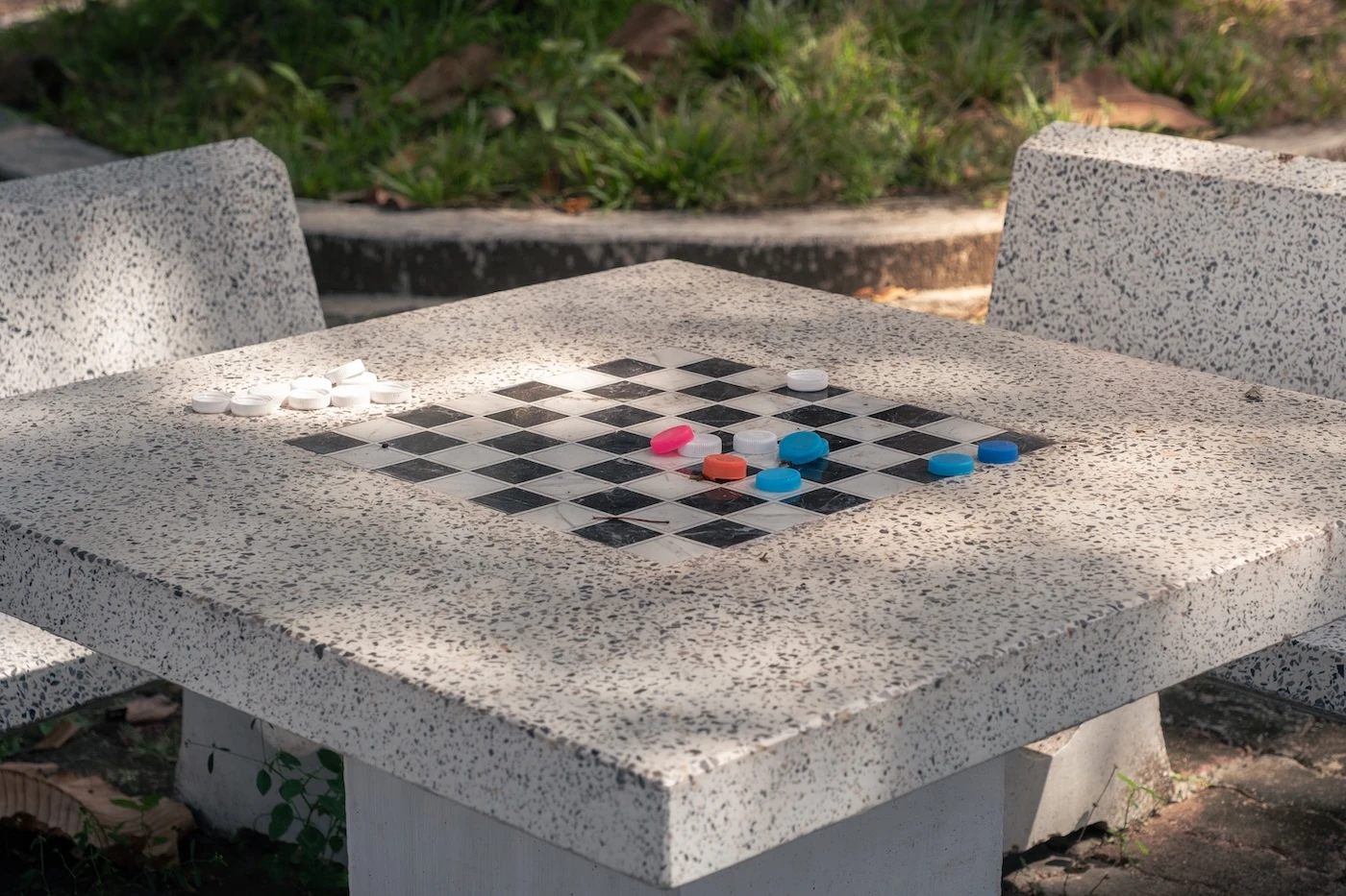 An outside stone table and chairs in the Wat Bang Thong Temple complex with a checkers board painted on the table surface and some coloured bottlecaps.